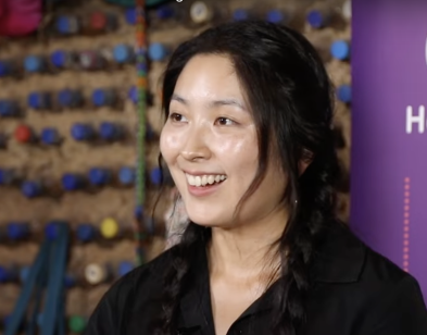 A woman with braided hair smiles in front of a colorful indoor climbing wall.